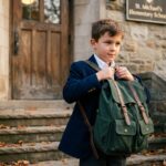 Jeune garçon portant un cartable vert structuré à l'entrée d'une école en pierre à l'automne